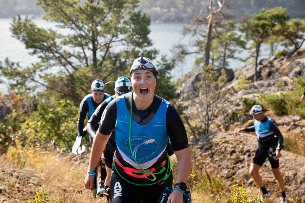A group of athletes in athletic gear, climbing a rocky path near a body of water, with one athlete laughing and appearing excited.