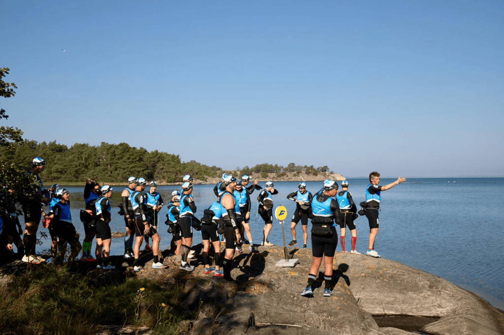 A group of athletes in blue and black gear gathering on a rock by the water, with trees in the background and a clear sky. One person is giving a speech while others listen attentively.