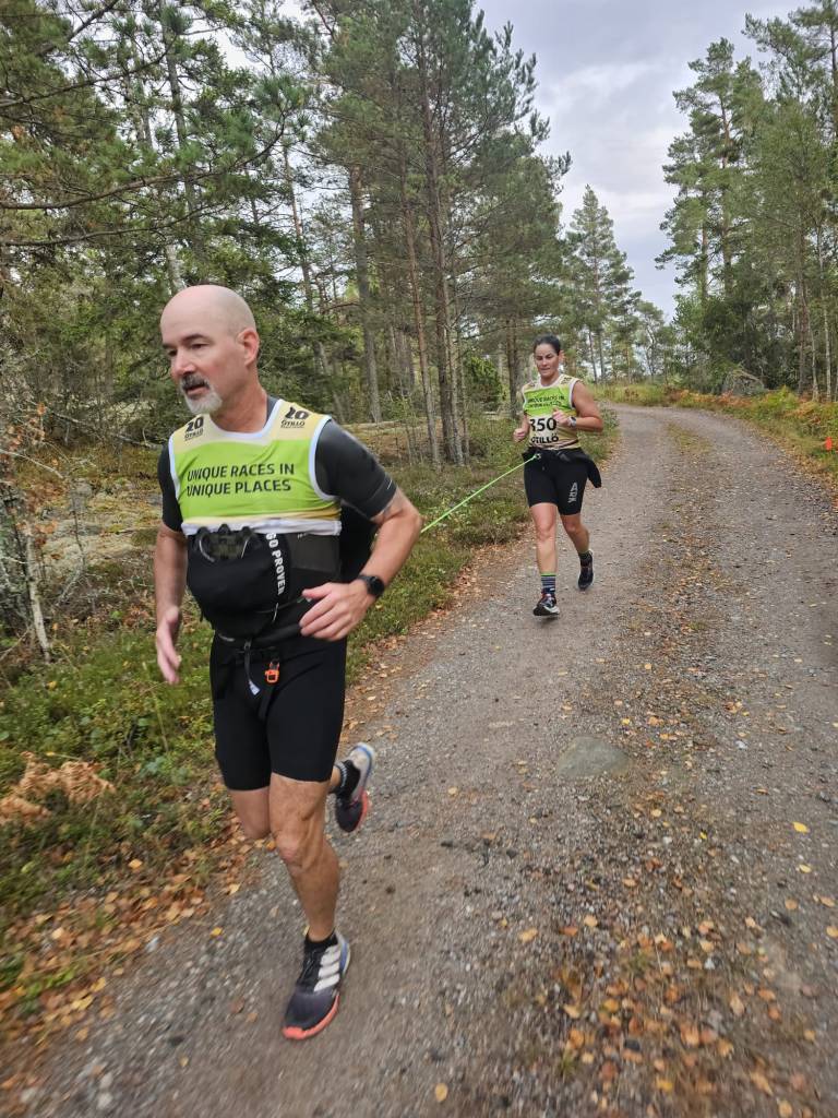 Two runners participating in a trail run through a forested path, wearing matching event shirts, with trees and foliage in the background.