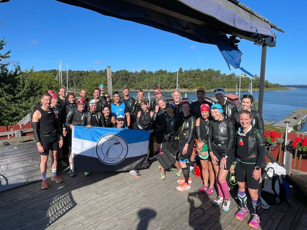 A group of swimmers in wetsuits poses together on a wooden deck with a waterfront view, holding an Envol Swimrun flag. The setting features a sunny blue sky, trees in the background, and boats in the water.