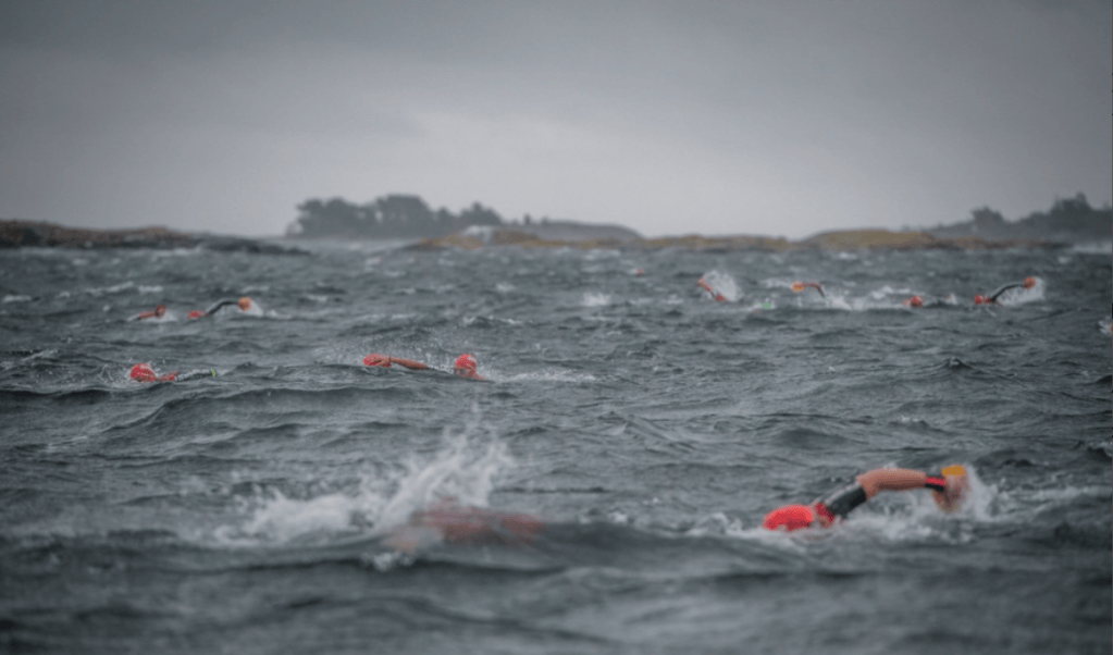 A group of swimmers wearing bright red caps competing in rough waters under a cloudy sky.
