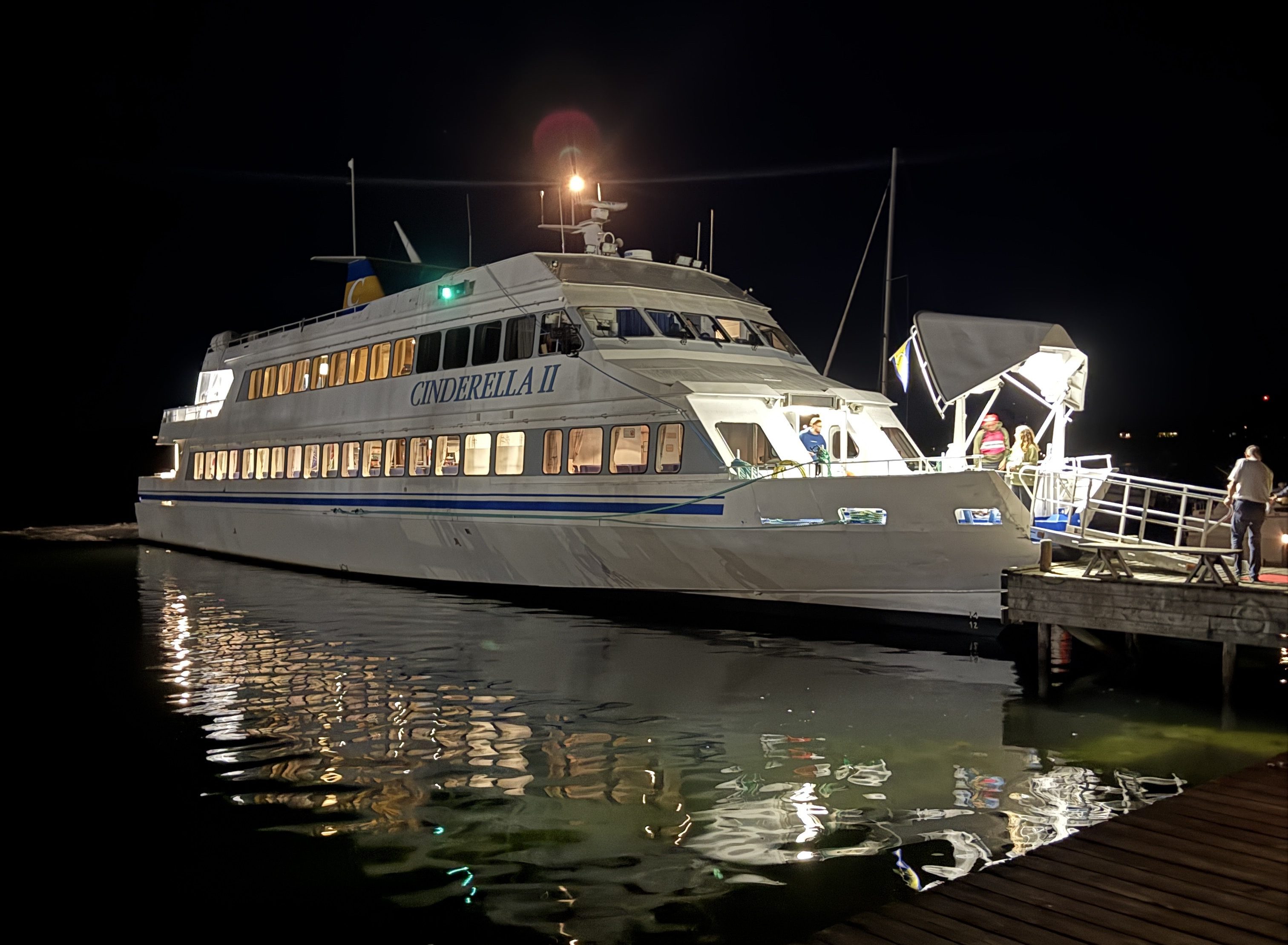A large white boat named Cinderella II docked at night, with lights illuminating the water and a gangway leading to the vessel.