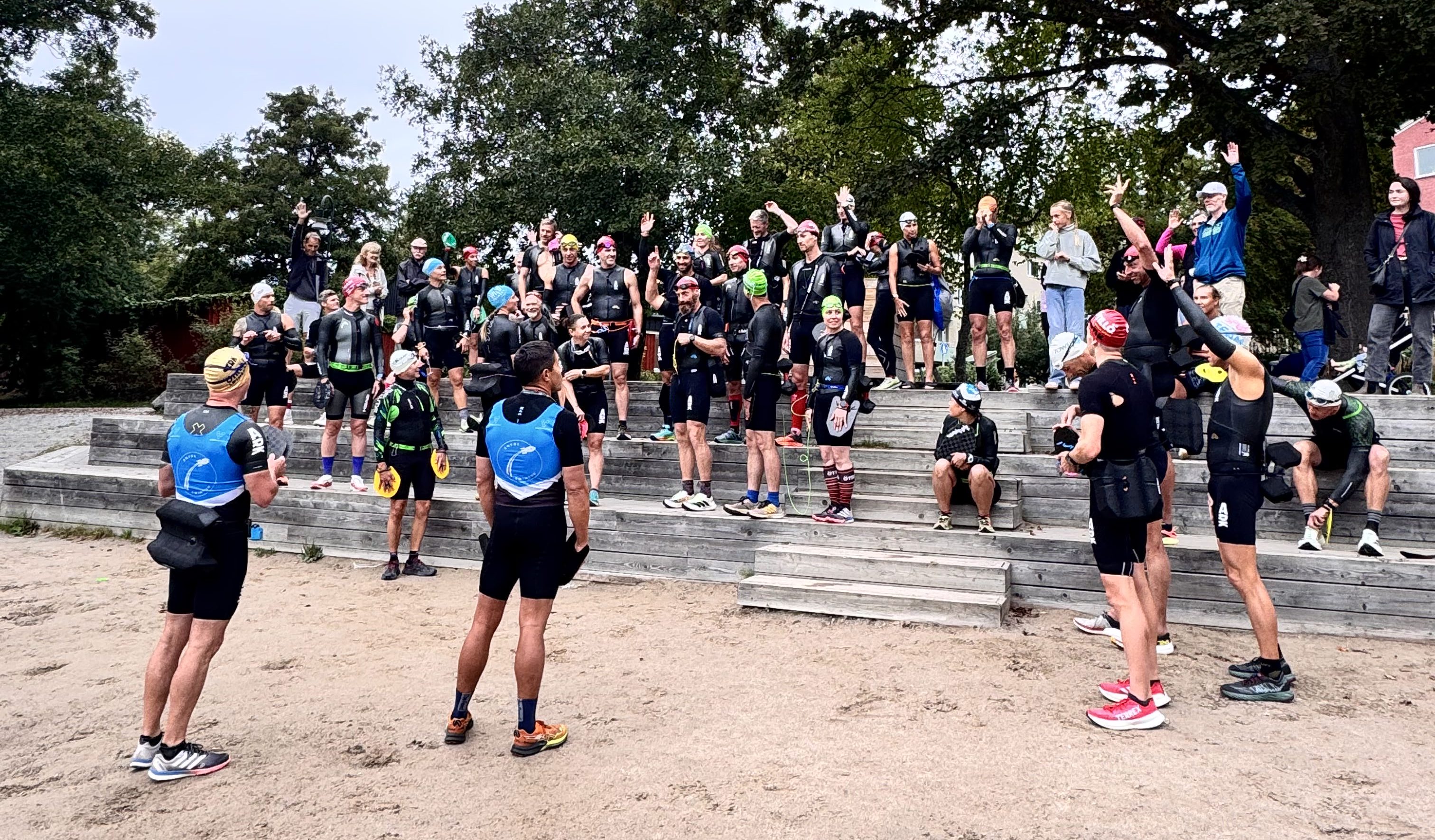 A large group of athletes in wetsuits and swim caps gathered on wooden steps, celebrating with raised hands, some sitting on the steps, while others pose for a photo.