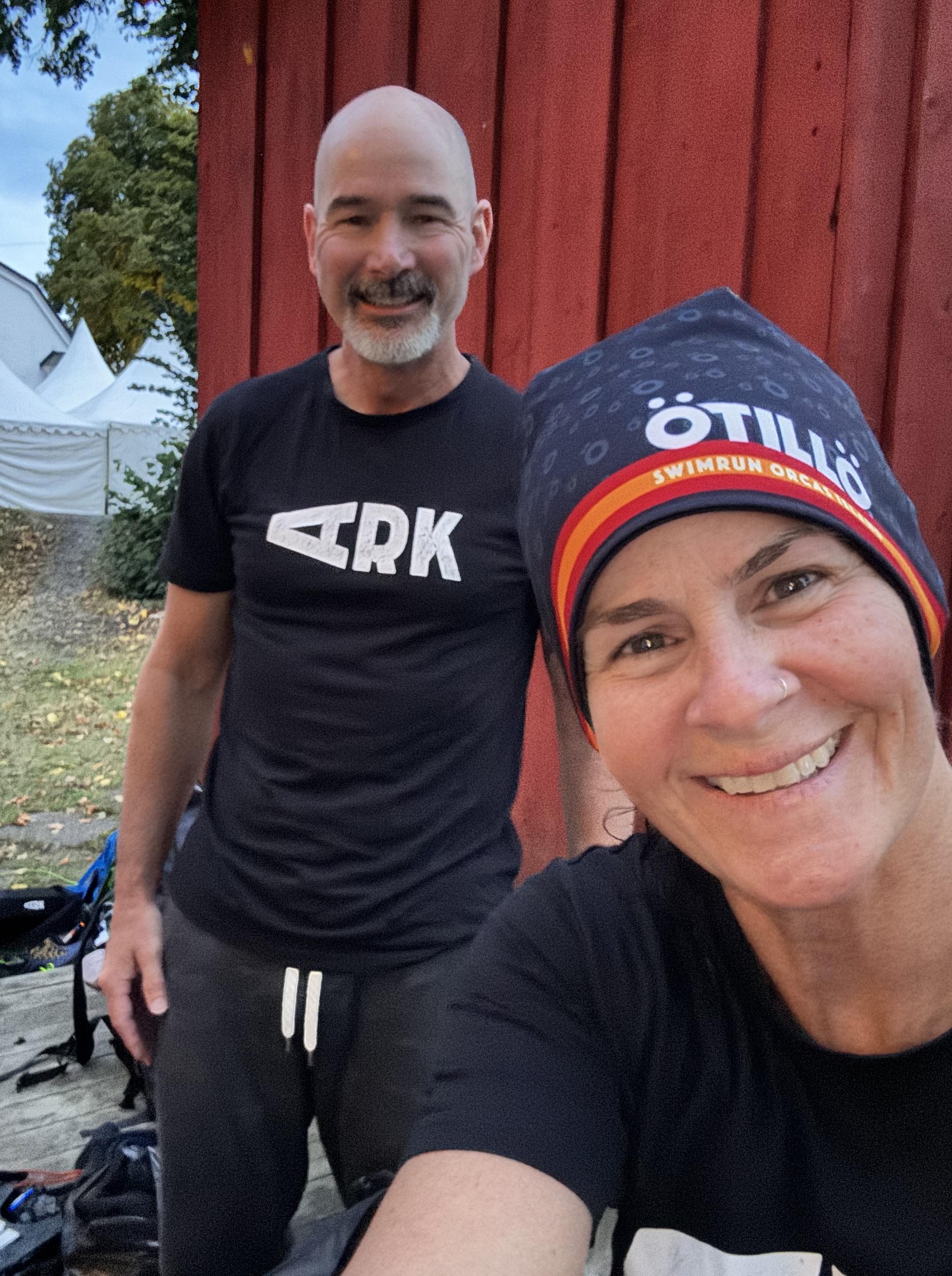 Two individuals posing for a selfie outdoors, one wearing a swim cap and the other in a black t-shirt with the word 'ARK'. They are in front of a red wooden wall.