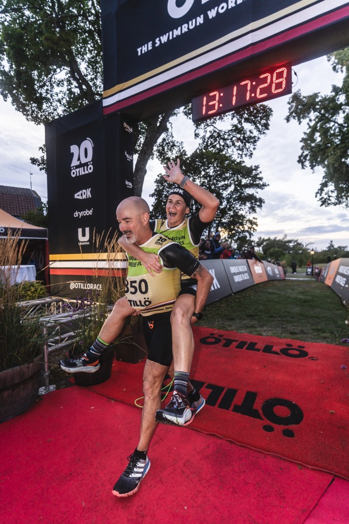 Two athletes celebrating at the finish line of a swimrun event, one is carrying the other on their back. A timer displays '13:17:28' above them.