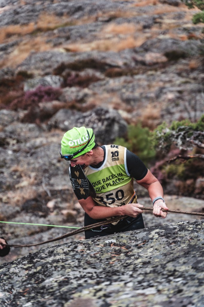 A climber in a green cap and black athletic gear navigates a rocky terrain, using a rope for support. They are wearing a numbered bib featuring '350' and focused on the ascent.