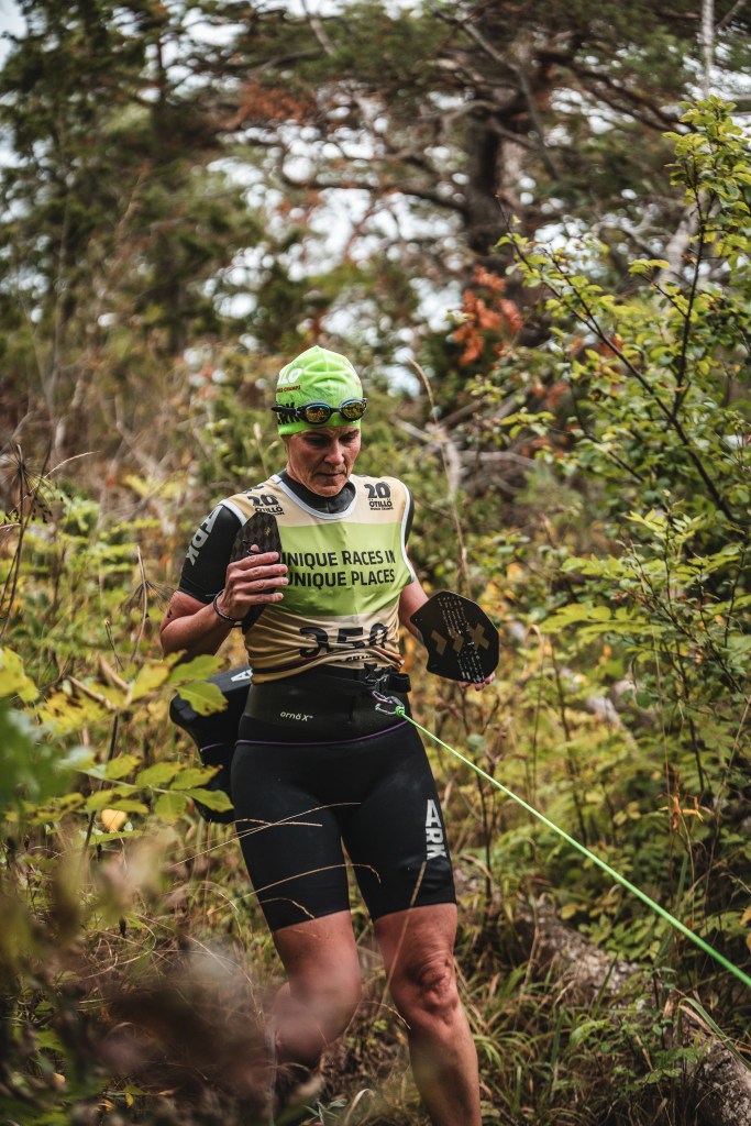 A female athlete wearing a green swim cap and goggles, holding a paddle, is navigating through a dense forest during a race.