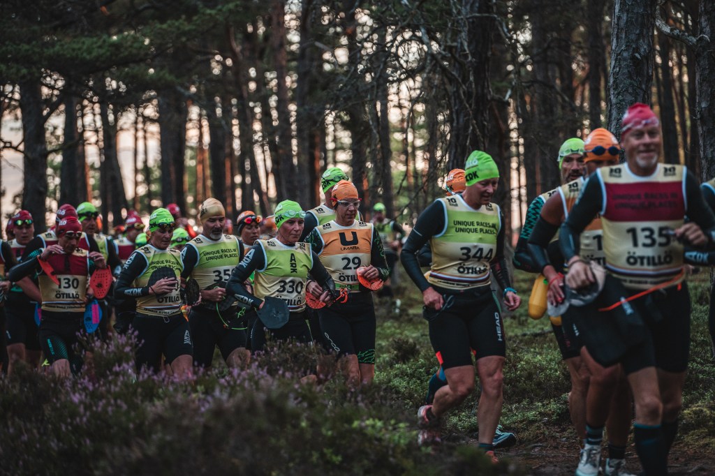 A group of athletes participating in an ÖTILLÖ swimrun event, wearing colorful swim caps and wetsuits, running through a forested area.