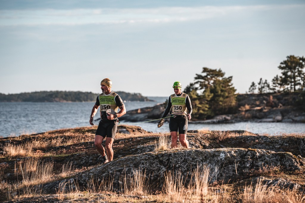 Two men wearing athletic gear and numbers on their shirts are participating in a running event along rocky terrain by the water.