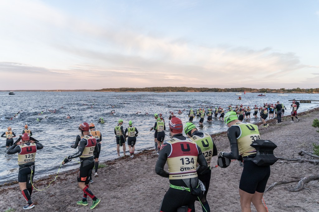 A group of triathletes in wetsuits preparing to enter the water for a swim leg of a competition, with several competitors already swimming in the lake, under a partially cloudy sky.