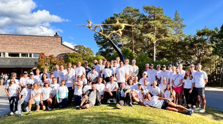 A large group of people in matching white t-shirts posing together outdoors in front of a modern building and a large sculpture surrounded by trees.