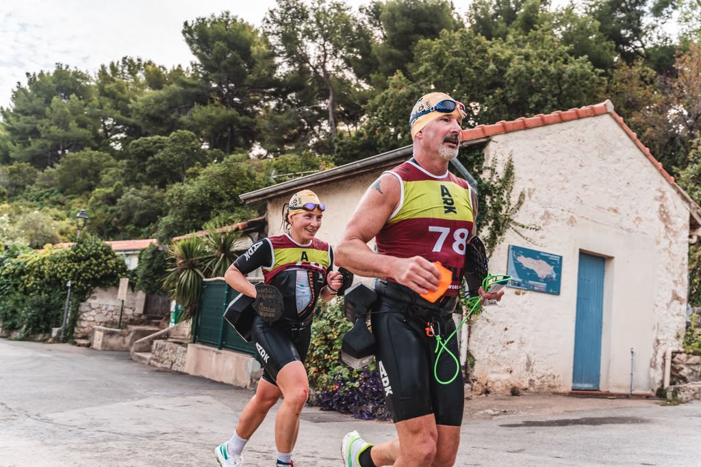 Two athletes running in a scenic outdoor setting, wearing swim gear and race numbers, with lush greenery and a house in the background.