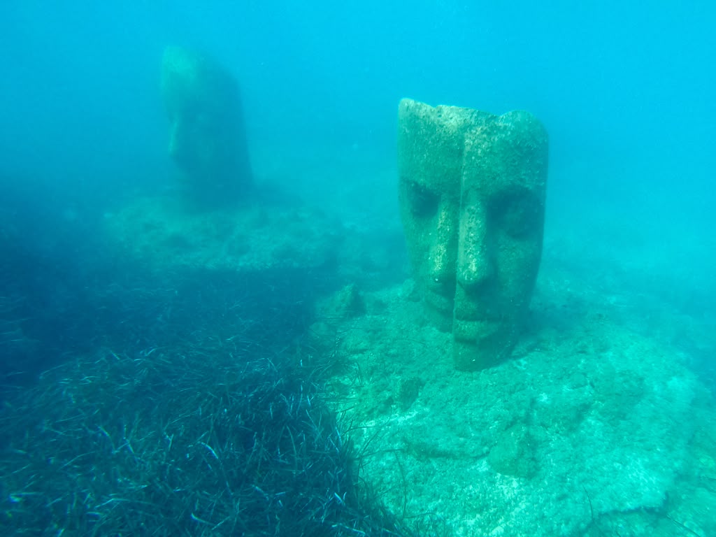 Underwater view of two ancient stone face sculptures partially submerged in sand and marine grass.