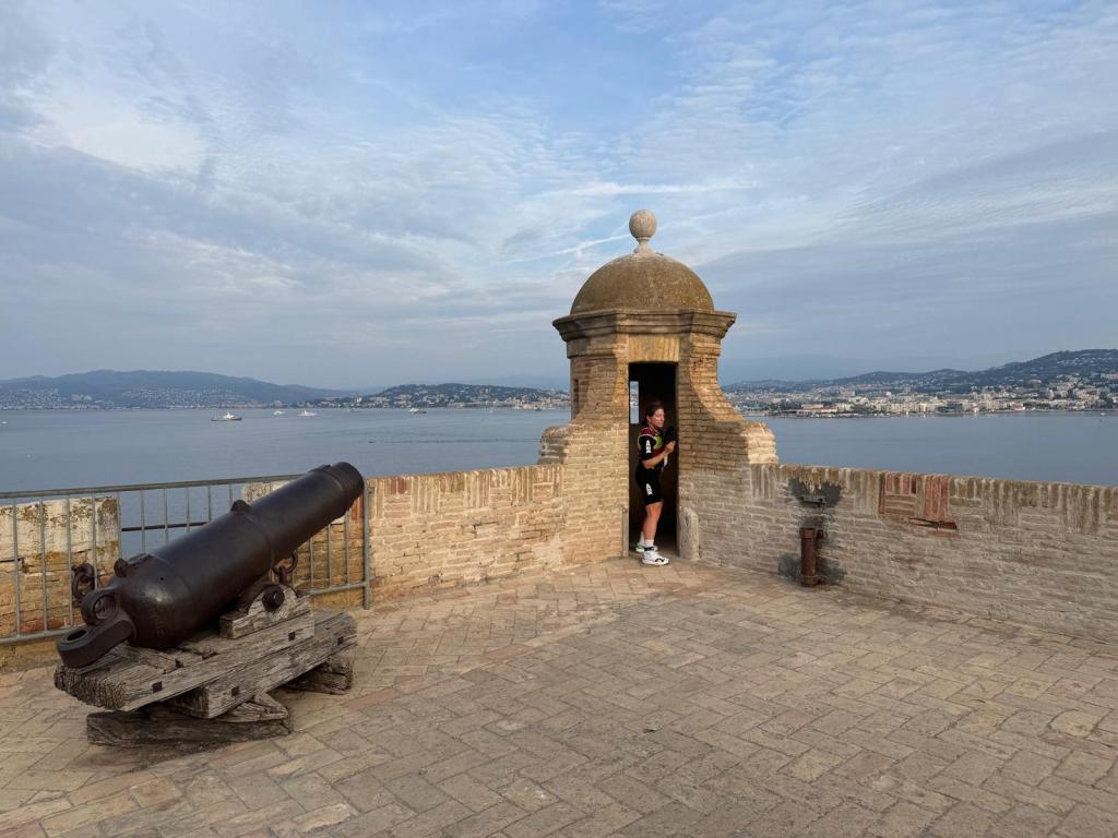 A person standing in a tower entrance on a coastal fortress, with a cannon in the foreground and a scenic view of the sea and distant hills.