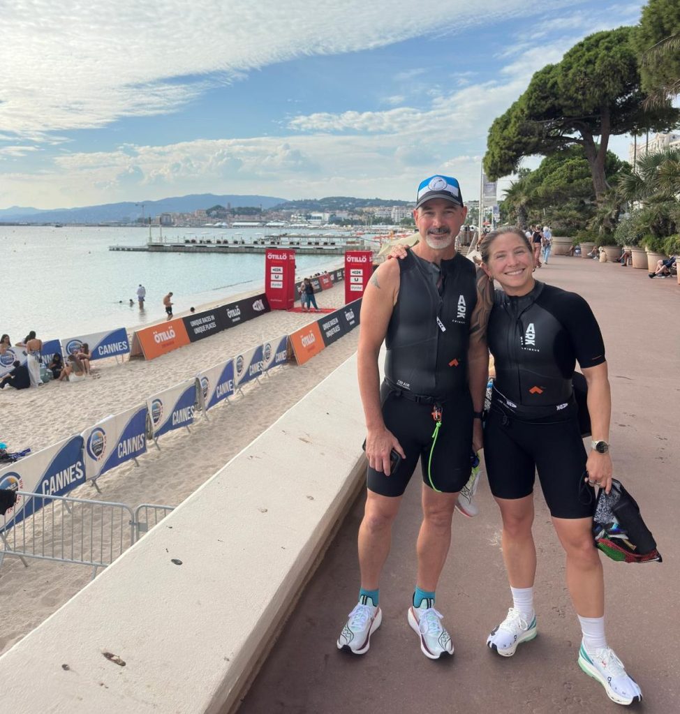 Two athletes in wetsuits smiling together at a beachside event in Cannes, with a scenic view of the ocean and coastline in the background.