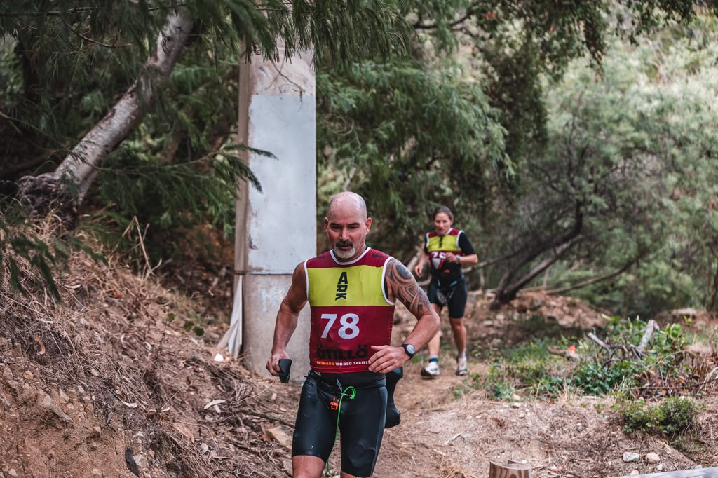 Two athletes running on a rugged outdoor trail, with one man in the foreground wearing a red and yellow jersey with the number 78 and a woman behind him in a similar outfit.