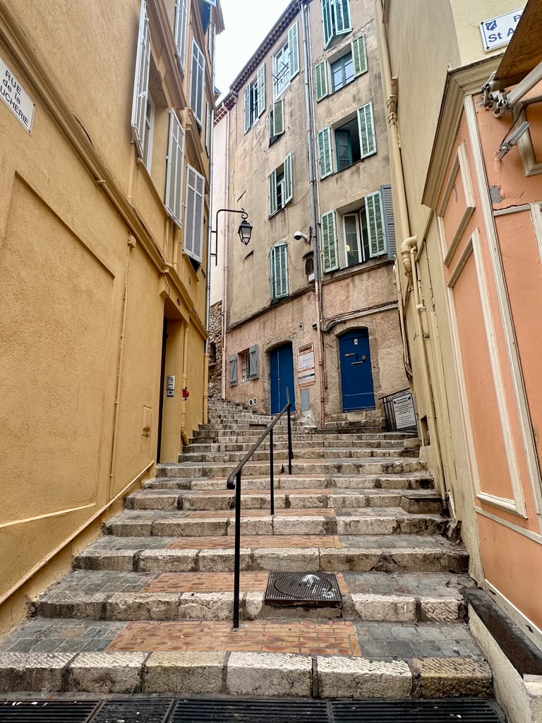 A narrow, cobblestone stairway winding between colorful buildings with shutters, leading upwards in a quaint urban setting.