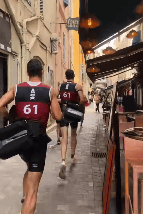 Two athletes in black and red running jerseys with numbers 61 and 62 jog through a narrow street lined with shops and cafes.