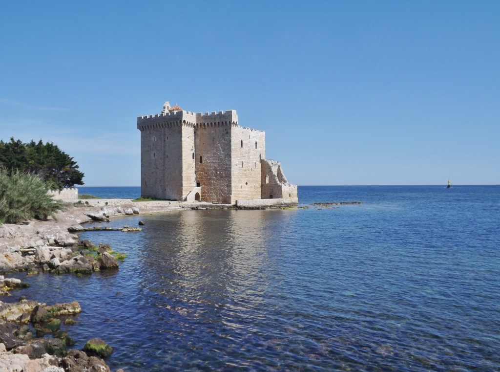 A historic stone castle standing on a rocky shore by the sea, surrounded by calm blue waters and green foliage under a clear blue sky.